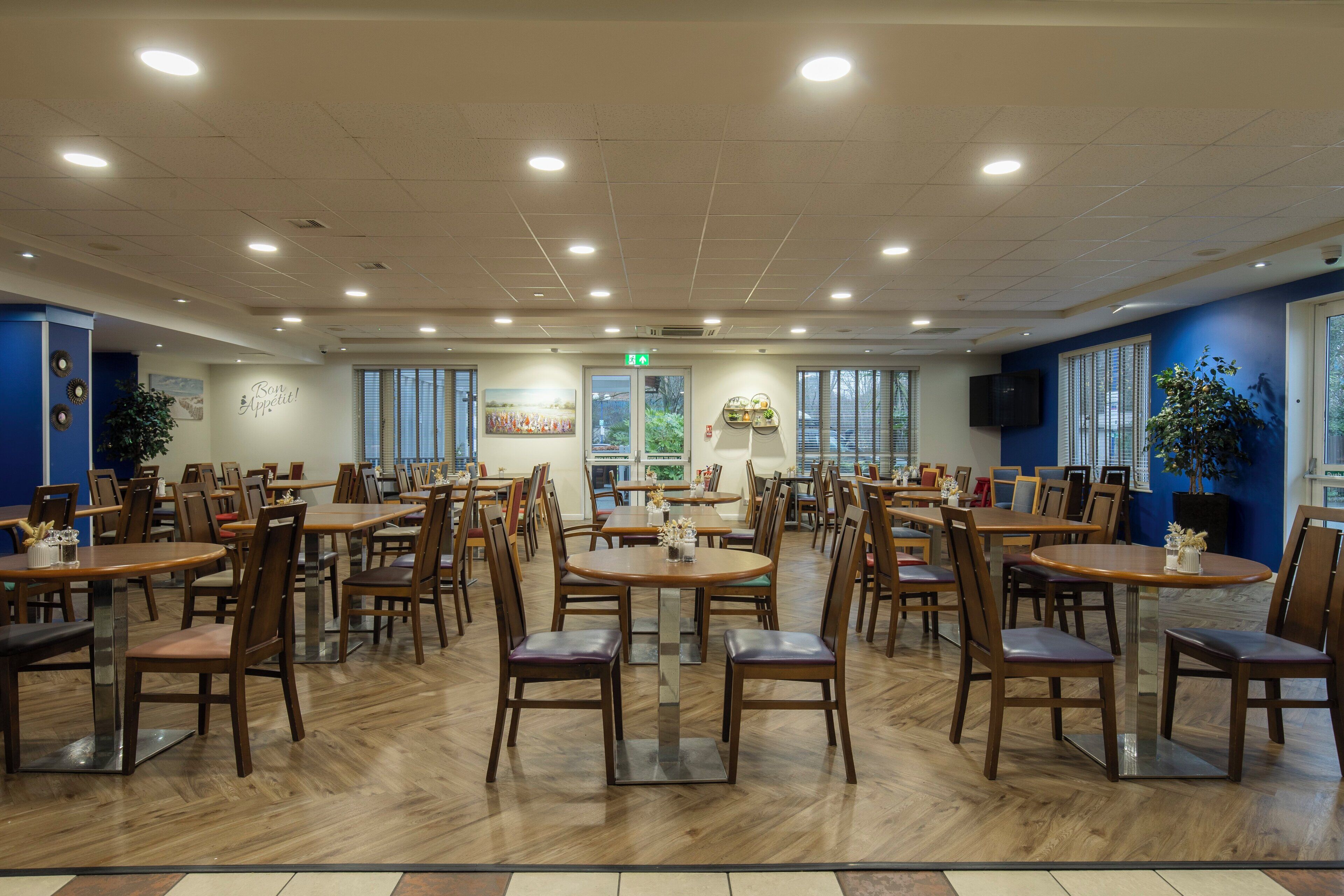 Un vaste espace dédié à la salle à manger, avec de nombreuses tables et chaises, un panneau de signalisation indiquant la sortie principale et une grande fenêtre laissant entrer la lumière naturelle.