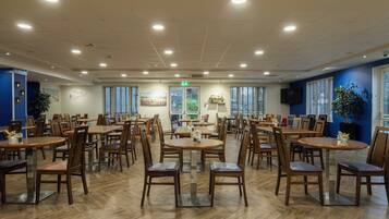 A spacious dining area with multiple tables and chairs, a central exit sign, and a large window providing natural light.