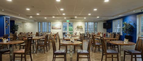 A spacious dining area with multiple tables and chairs, a central exit sign, and a large window providing natural light.