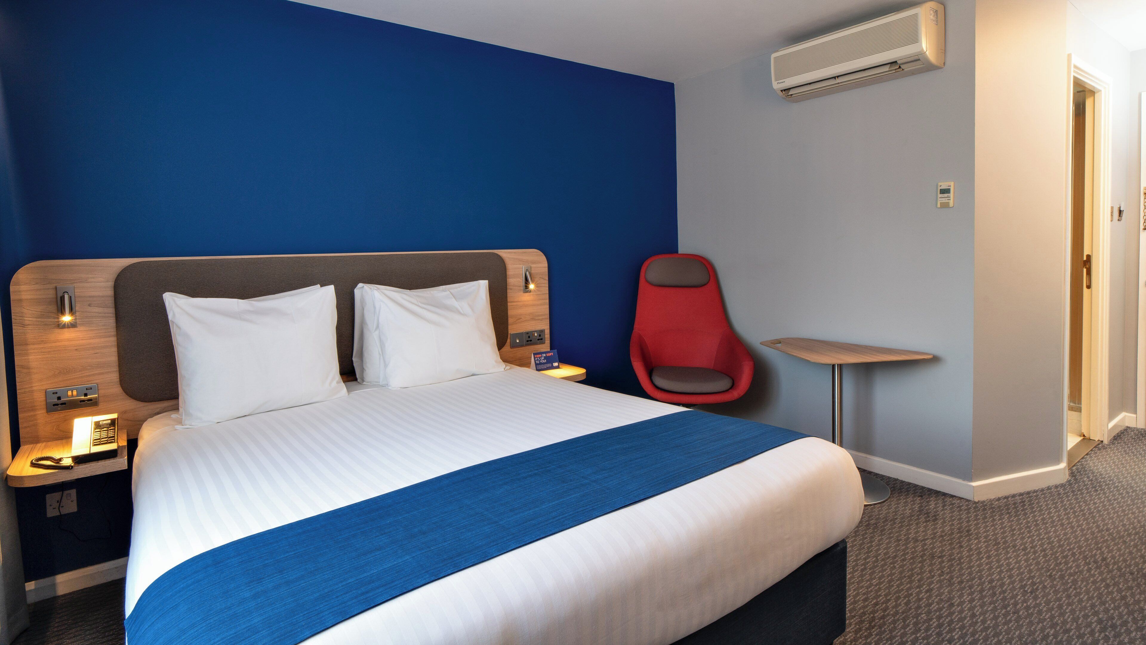 Una camera d'albergo con un letto grande, un comodino, una sedia rossa, un tavolino e un condizionatore d'aria a parete.