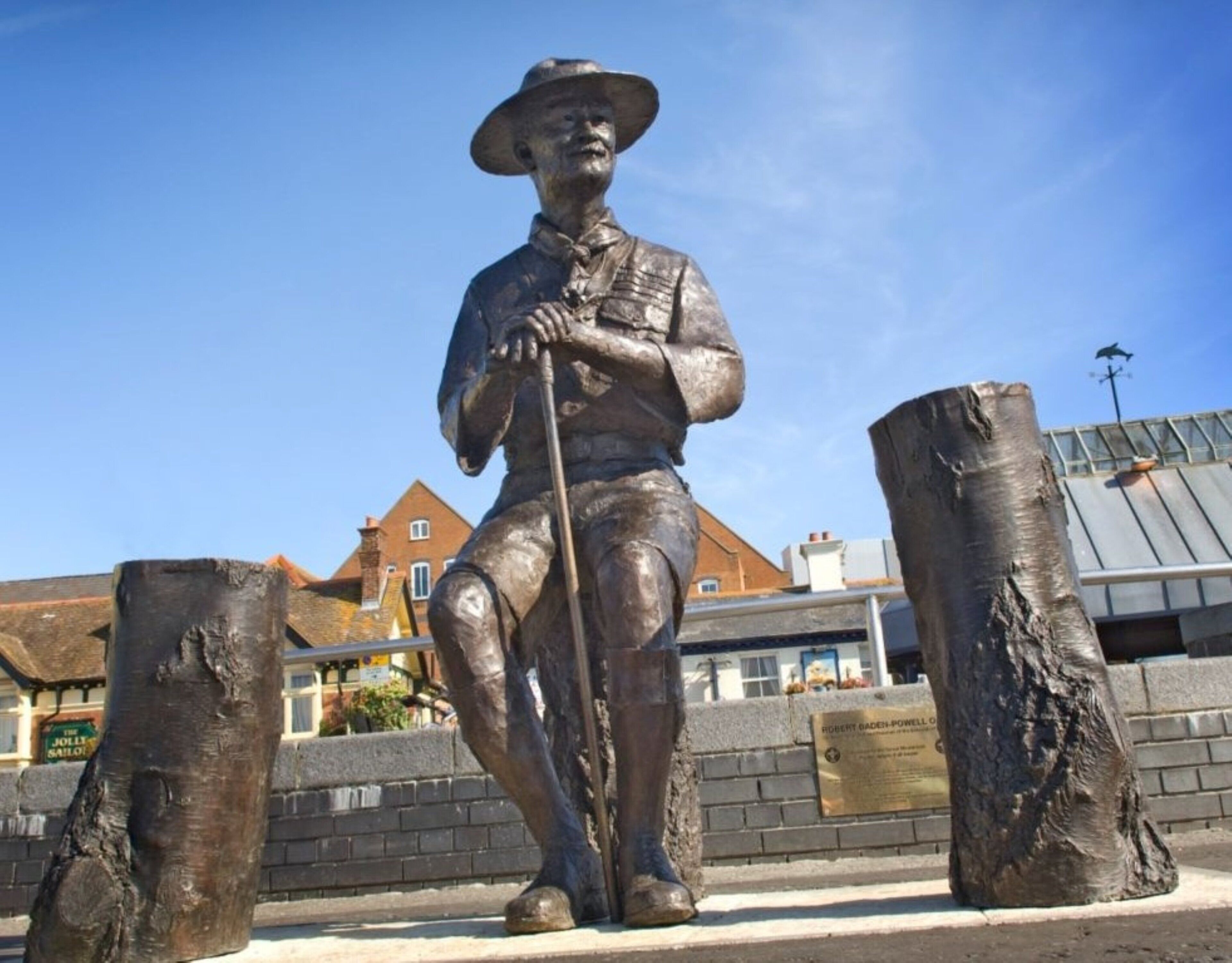 A bronze statue of a man in uniform with a hat and cane, standing on a paved area with a brick wall and buildings in the background.
