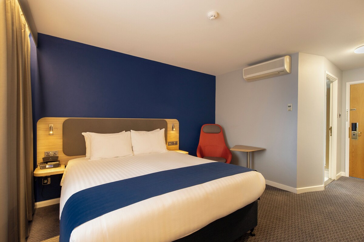 A hotel room with a large bed, a nightstand, a red chair, a small table, and an air conditioning unit.