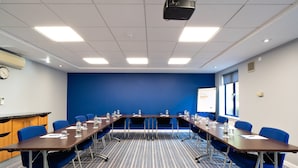 Une salle de conférence avec une longue table, des chaises bleues et un écran de projection.