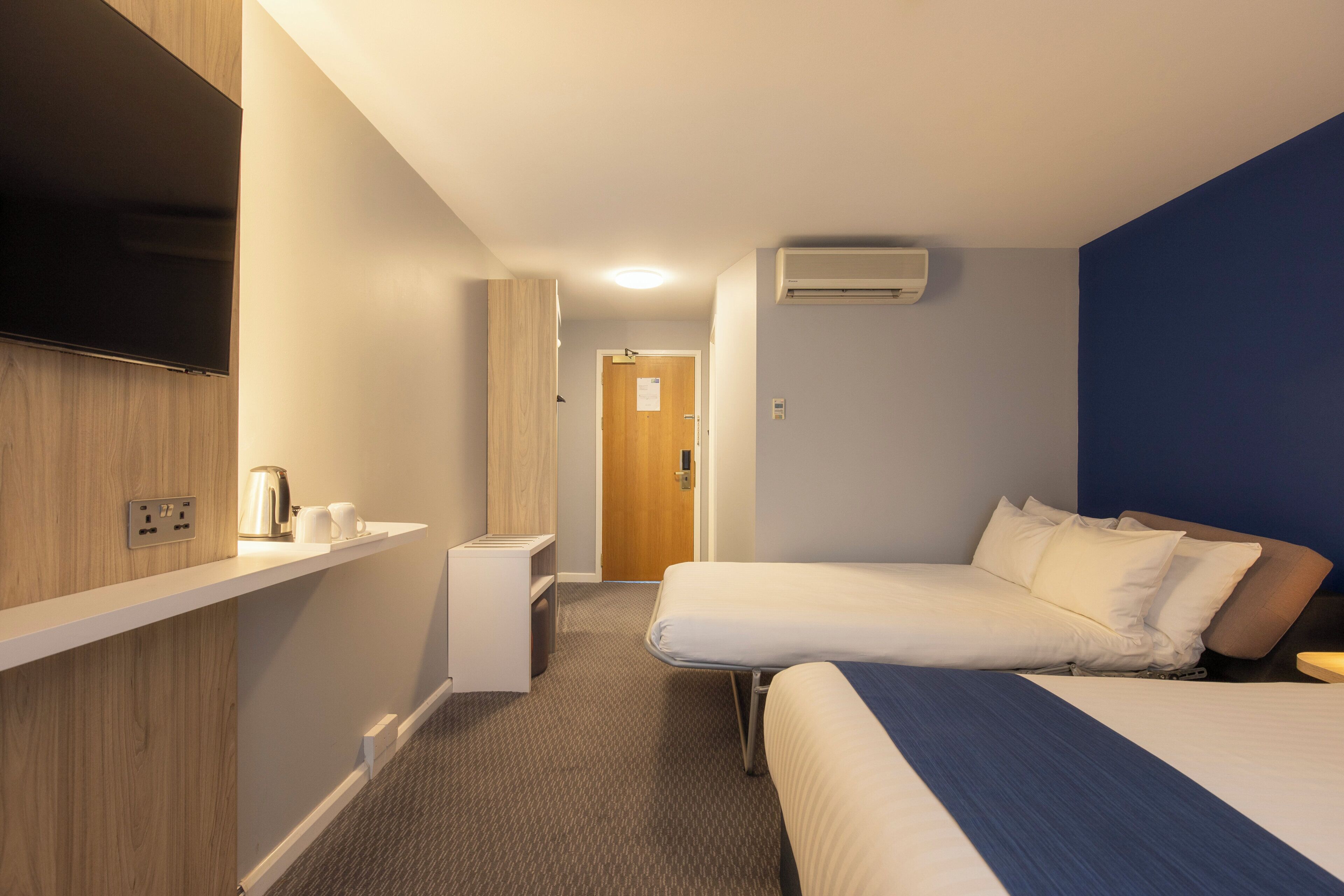 Una habitación de hotel con dos camas, un televisor de pantalla plana en la pared, una mesita de noche pequeña y un enchufe.