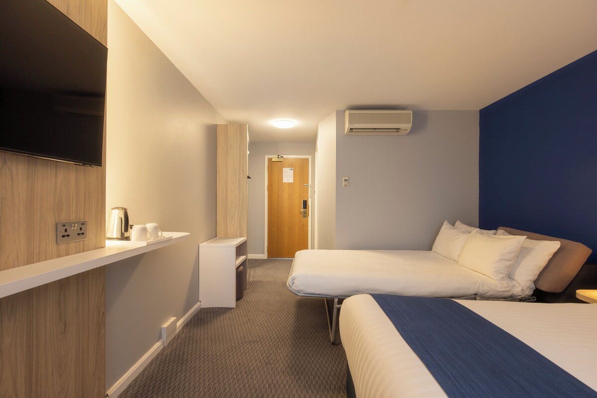 A hotel room with two beds, a flat-screen TV mounted on the wall, a small bedside table, and a power outlet.