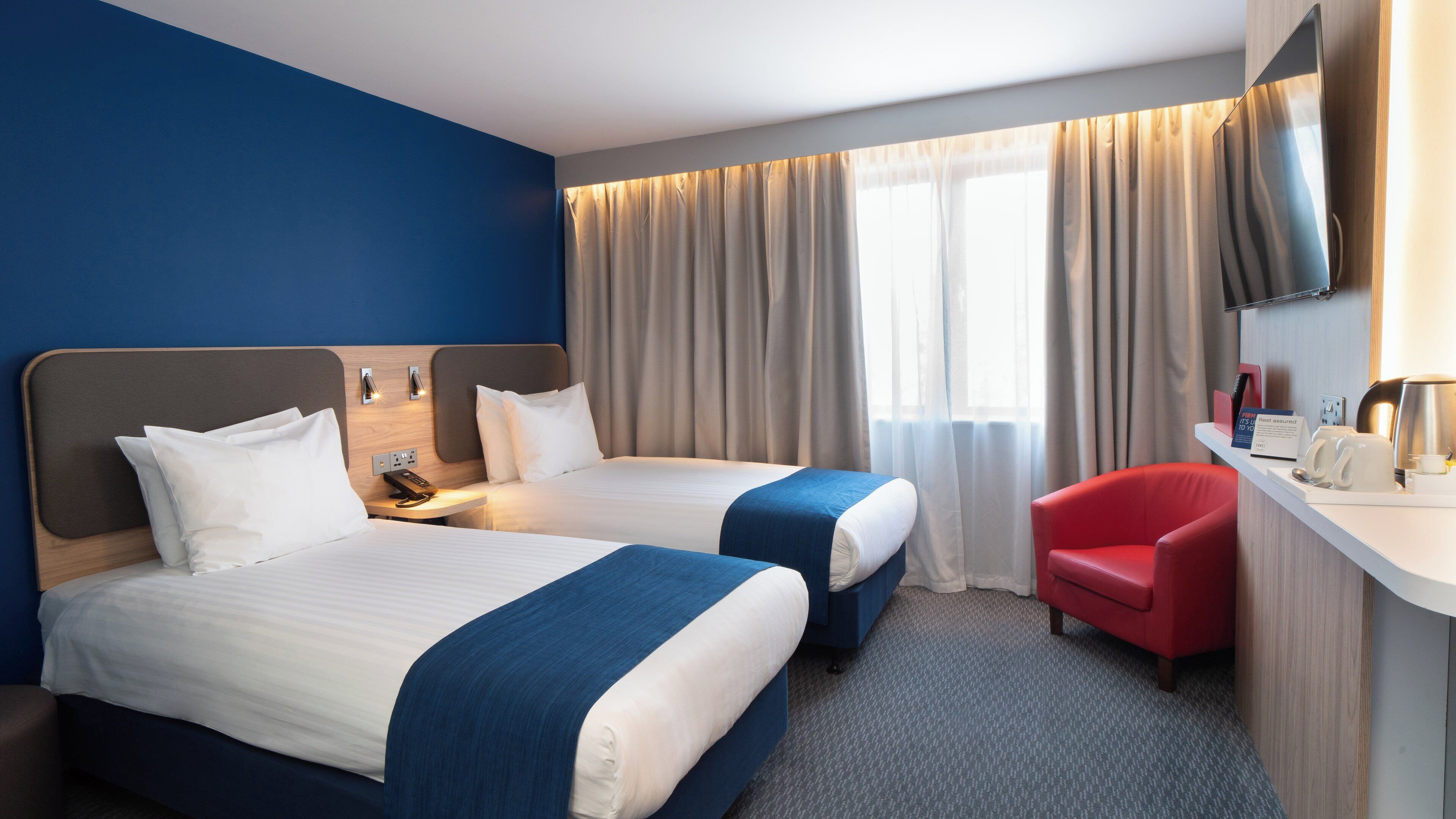 A hotel room with two beds, a desk with a red chair, a flat-screen TV, and a window with curtains.