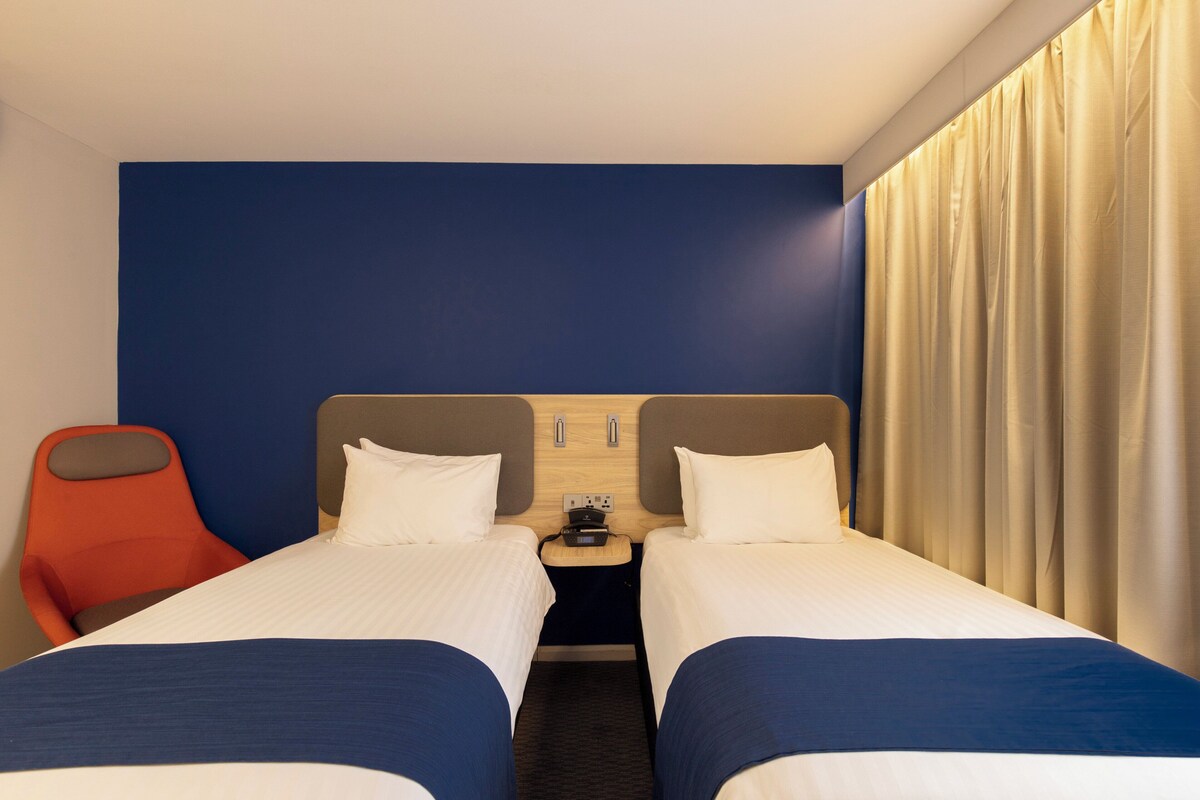 A hotel room with two beds, a nightstand, a red chair, and a curtain.
