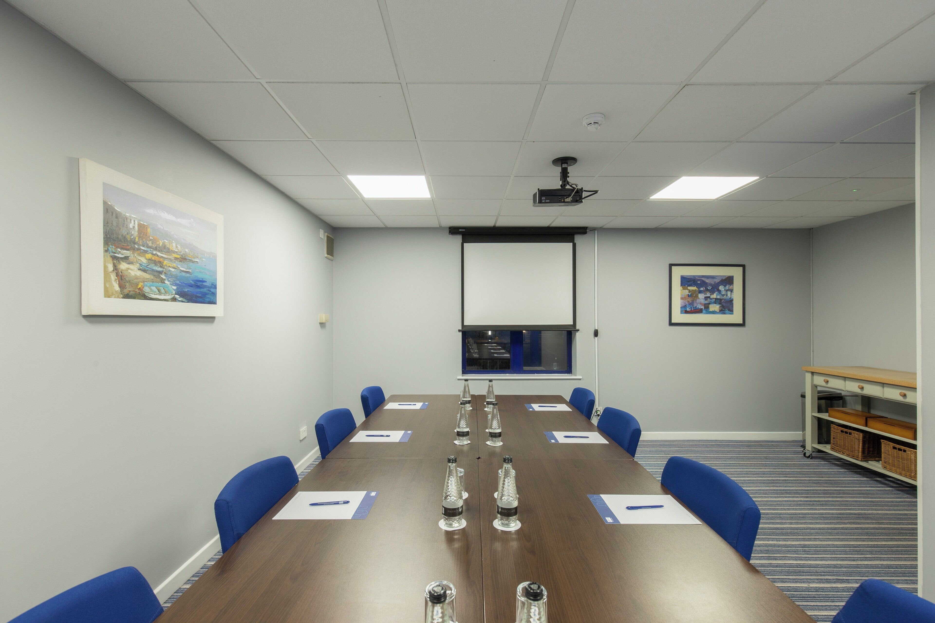 A conference room with a long table, blue chairs, water bottles, and a projector screen.