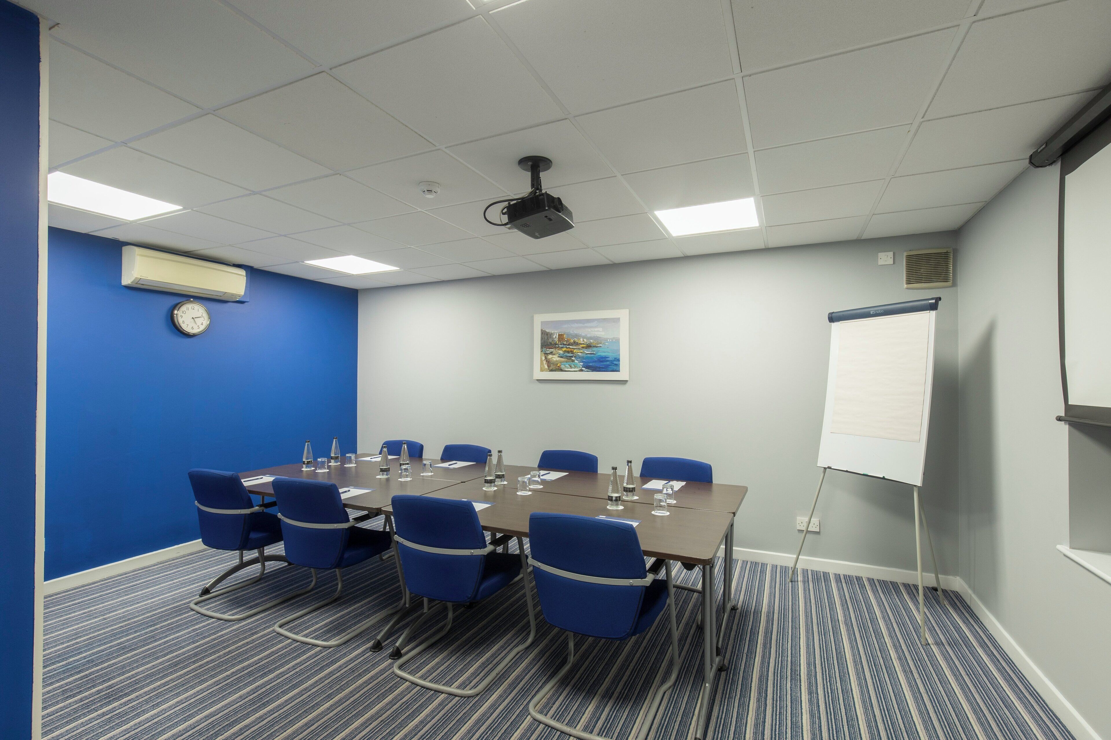 A conference room with a large table, blue chairs, a projector, a whiteboard, and a clock on the wall.