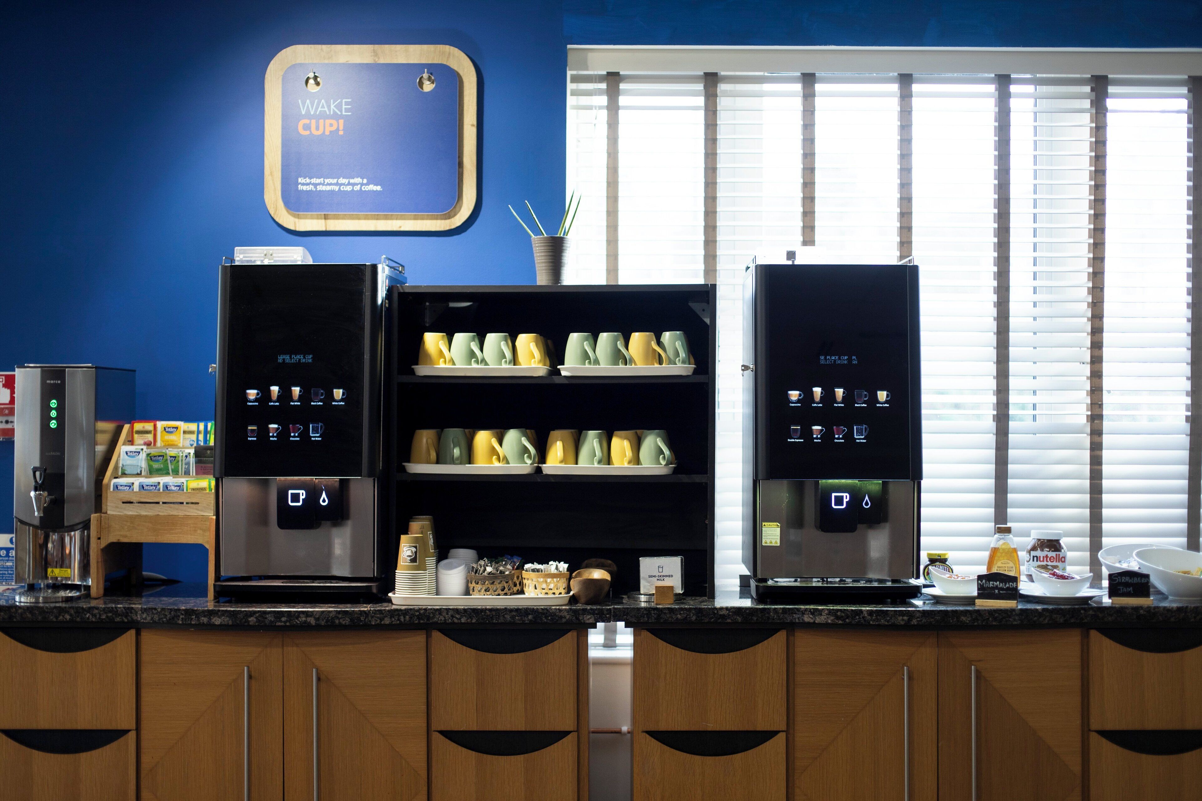 A coffee station with two coffee machines, a sign above them, and a window with blinds in the background.