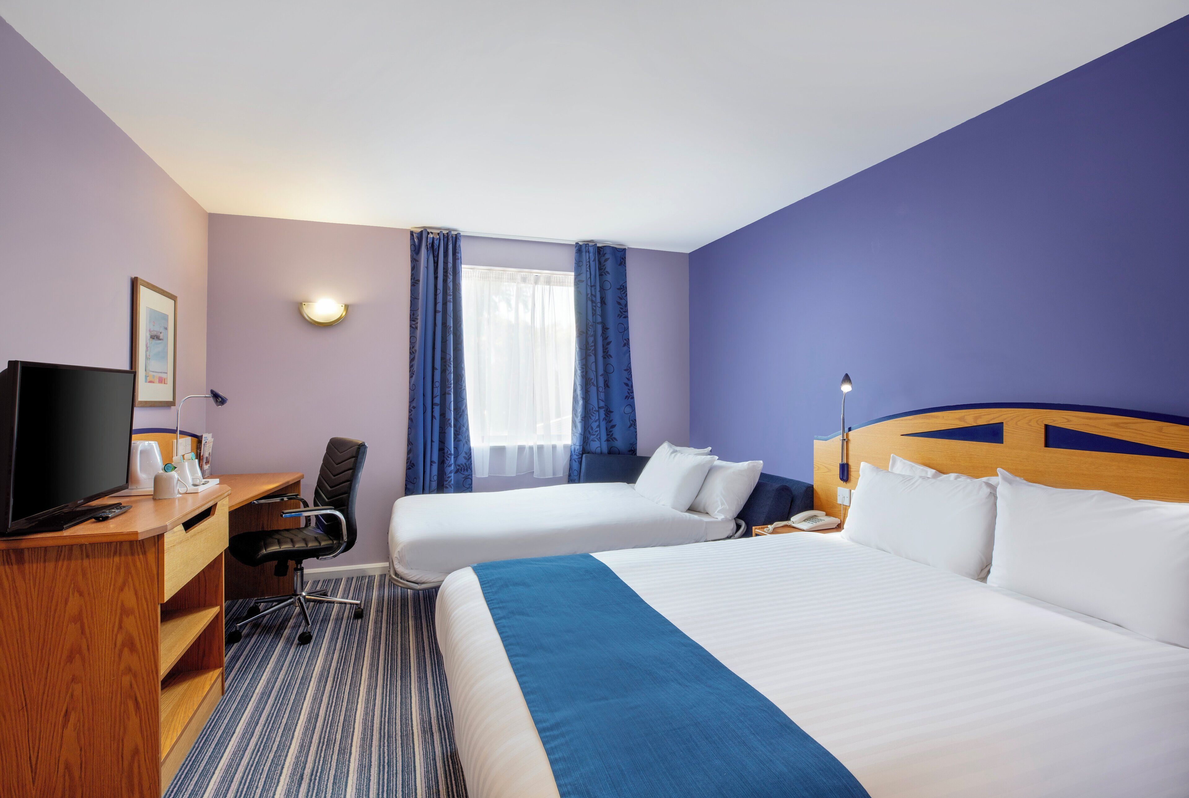 A hotel room with a bed, a desk with a TV, a chair, and a window with curtains.