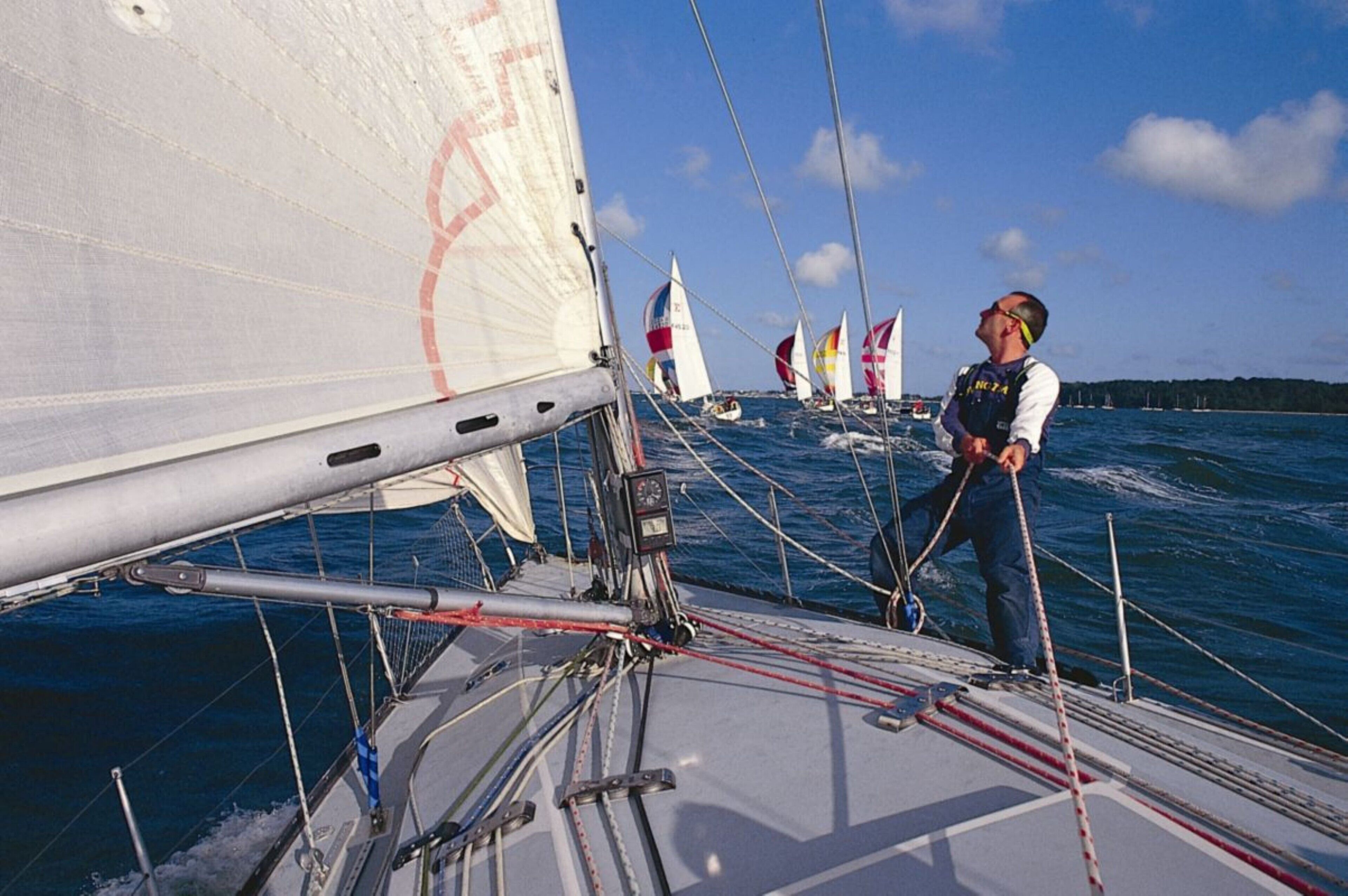 A sailboat with multiple sails, a person standing on the deck, and other sailboats in the background.