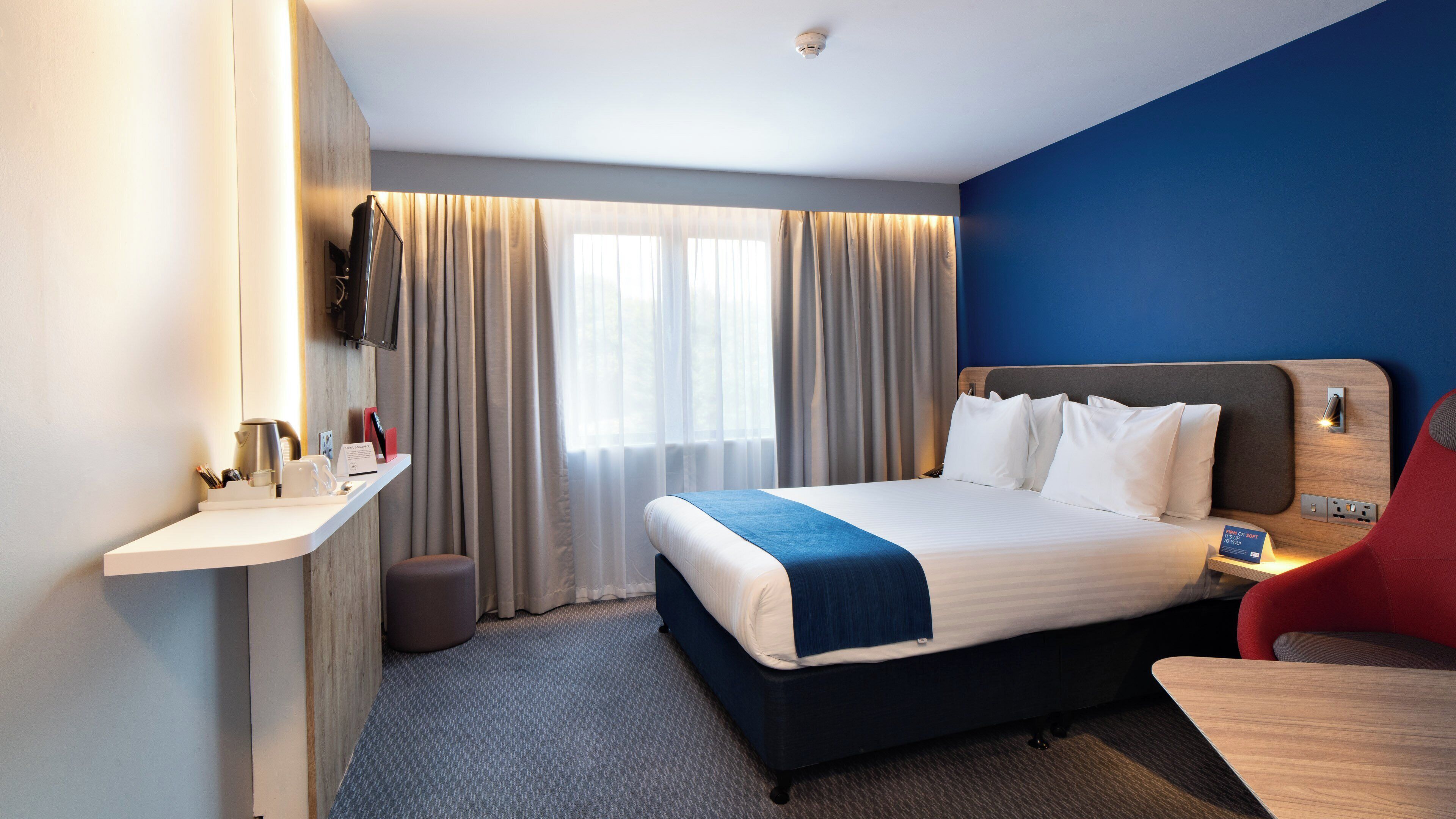 Ein Hotelzimmer mit einem Bett, einem Schreibtisch, einem Sessel, einem an der Wand befestigten Fernseher und einem Fenster mit Vorhängen.