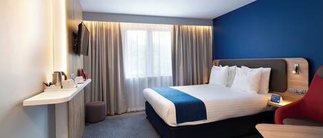 Ein Hotelzimmer mit einem Bett, einem Schreibtisch, einem Sessel, einem an der Wand befestigten Fernseher und einem Fenster mit Vorhängen.
