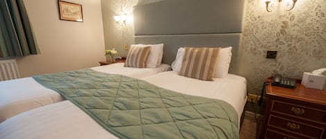 A hotel room with two beds, a green quilt, a nightstand with a phone, and a wall-mounted light fixture.