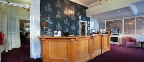 A reception area with a dark patterned wall, a wooden reception desk, a chandelier, and a brick wall with windows.