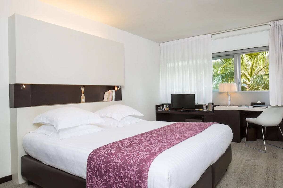 A hotel room with a bed, a desk with a computer, a chair, and a window with curtains.