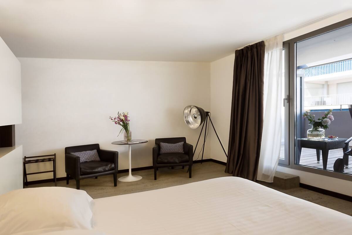 A hotel room with a large bed, two armchairs, a small round table with a vase of flowers, a floor lamp, and a view of a balcony with outdoor seating.