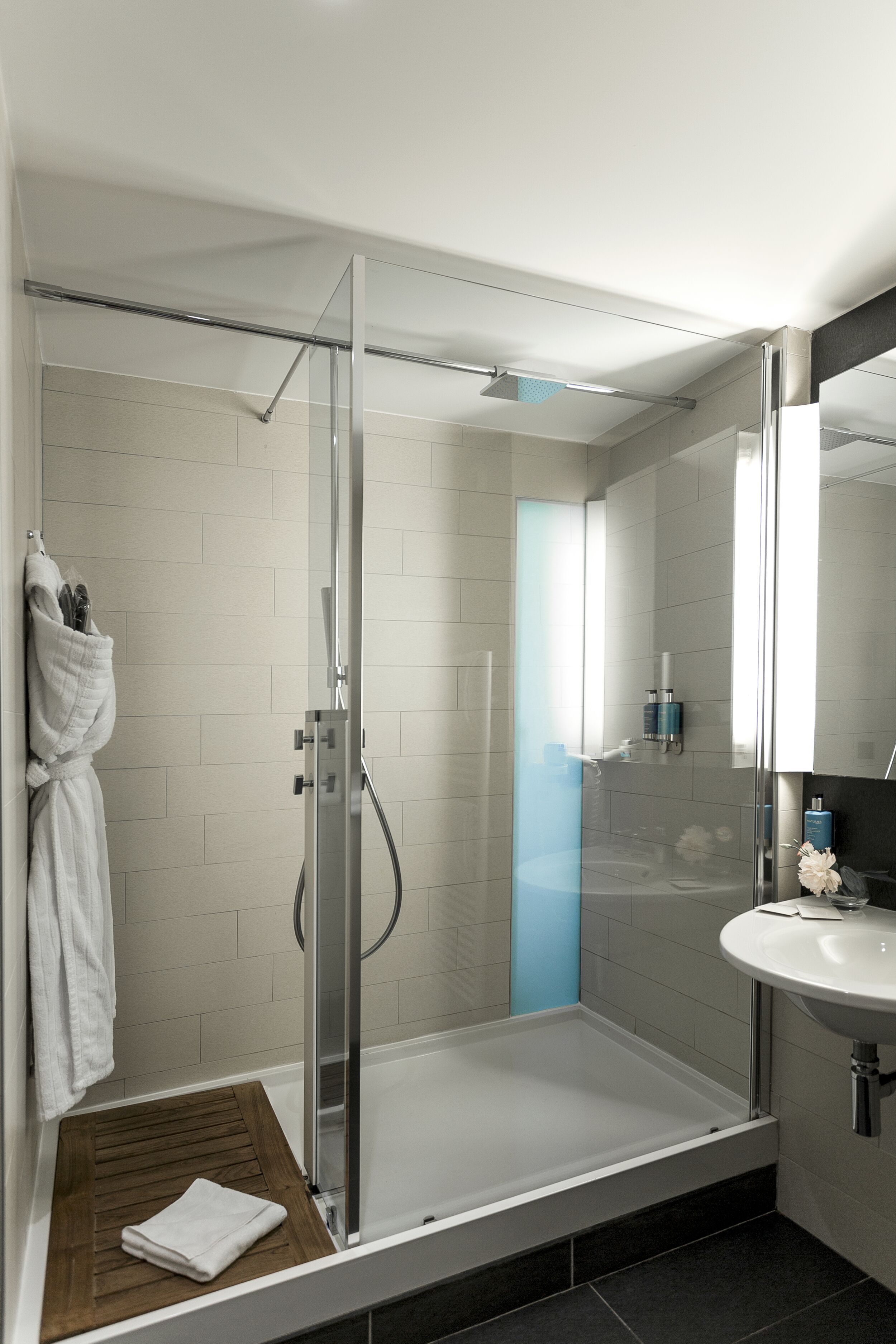 A modern bathroom with a glass-enclosed shower, a wooden bath mat, a white sink, and a mirror.