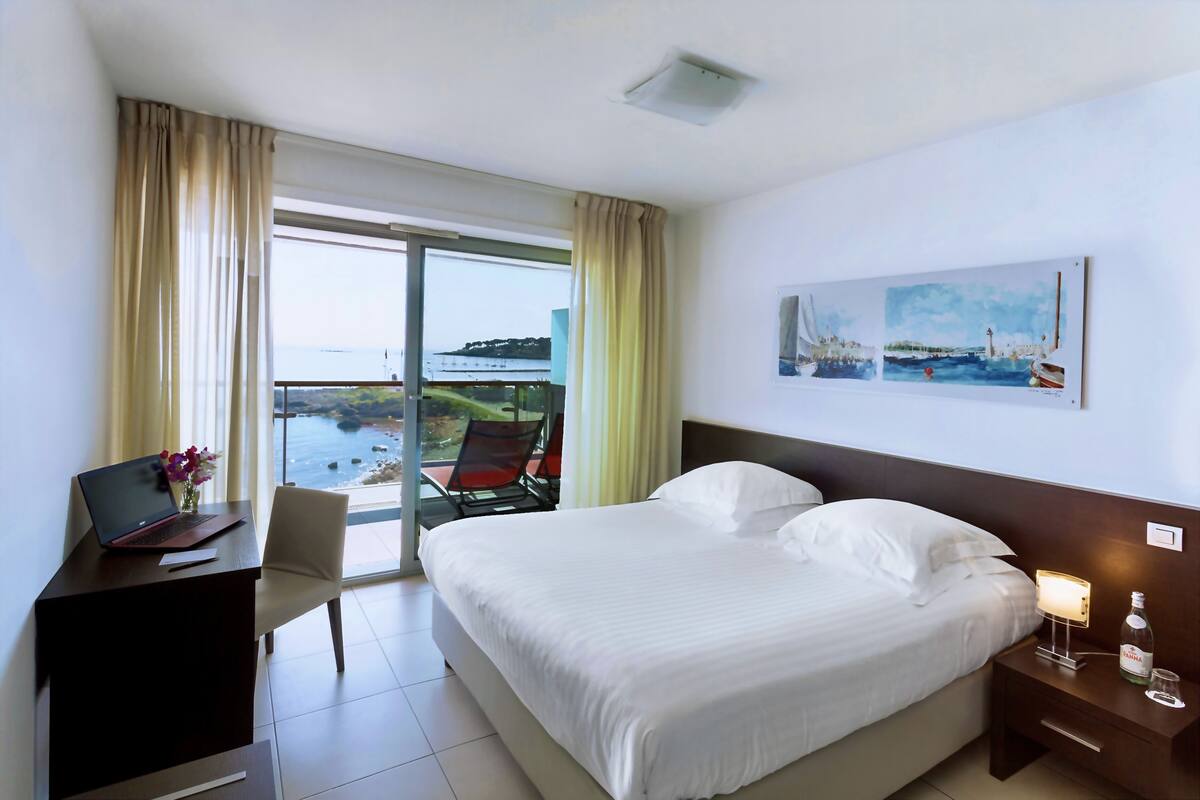 A hotel room with a bed, a desk with a laptop, a chair, a nightstand with a lamp, and a painting on the wall.
