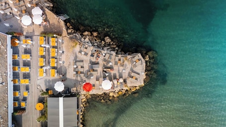An aerial view of a coastal resort with multiple sun loungers and umbrellas, a wooden deck, and a rocky shoreline.. Royal Antibes