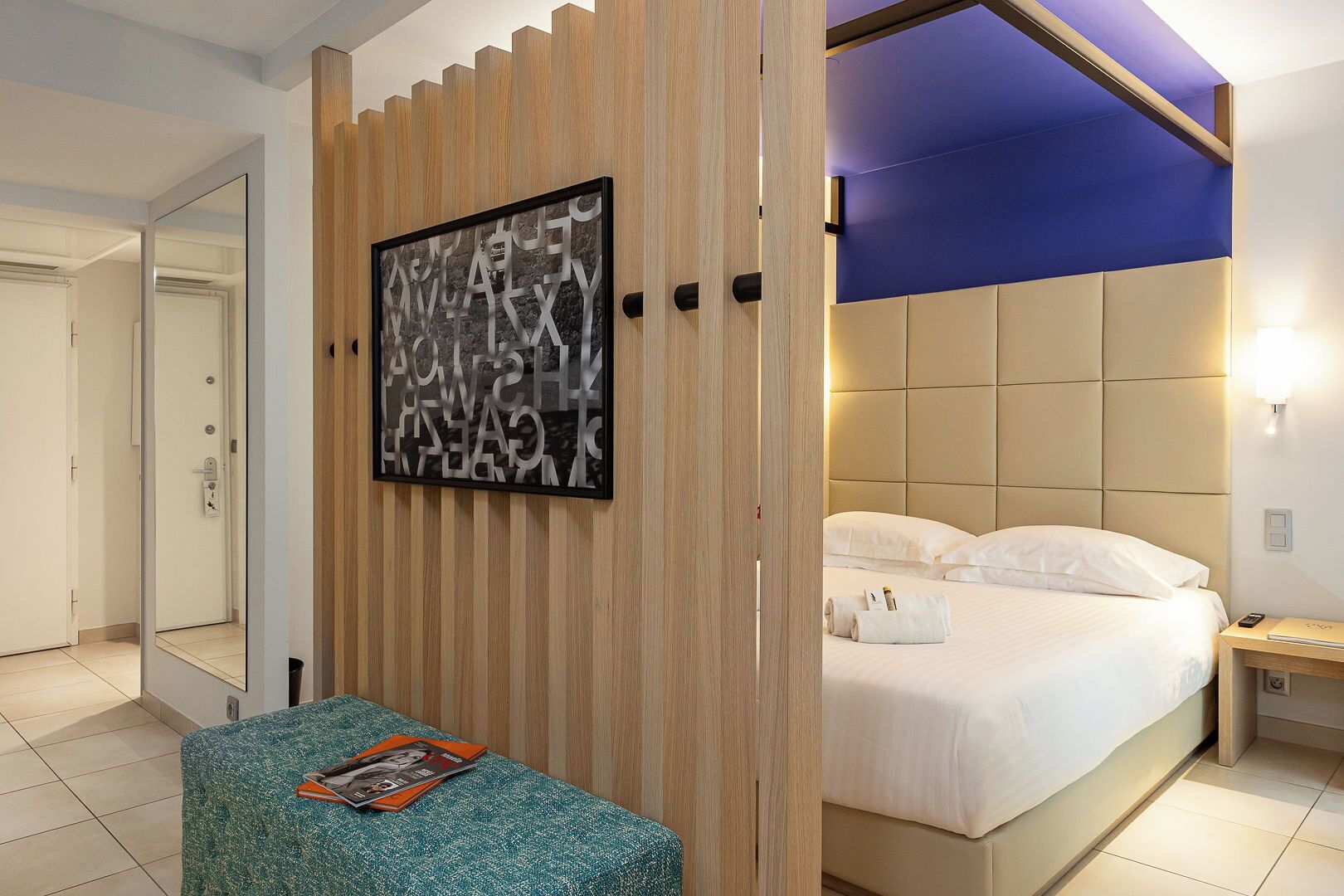 A hotel room with a bed, a bedside table with a lamp, a wall-mounted artwork, and a door leading to another room.
