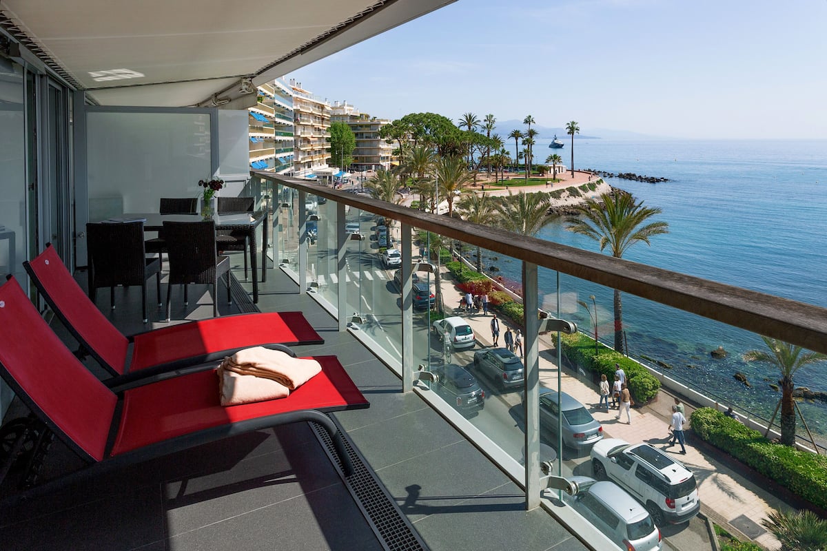 A balcony with red chairs, a table, and a view of the sea and buildings.