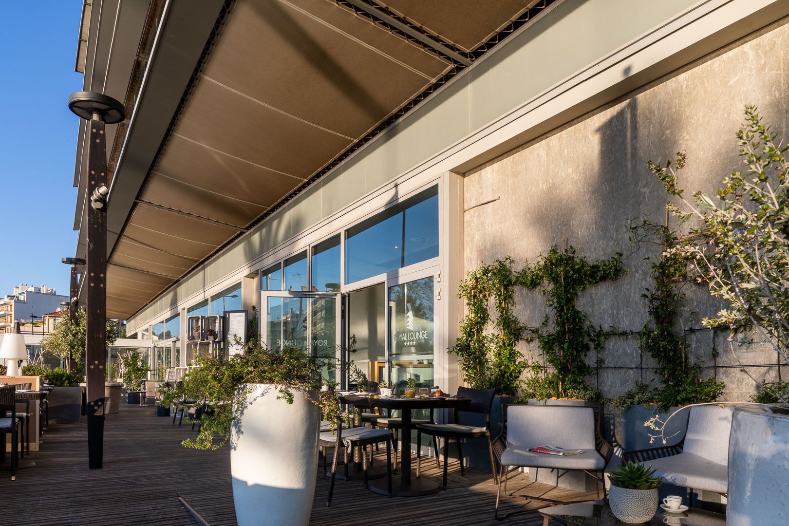 Uma área externa com mesas e cadeiras, uma grande planta em vaso e um edifício com janelas amplas e uma placa com a inscrição 'ROYAL LOUNGE'.