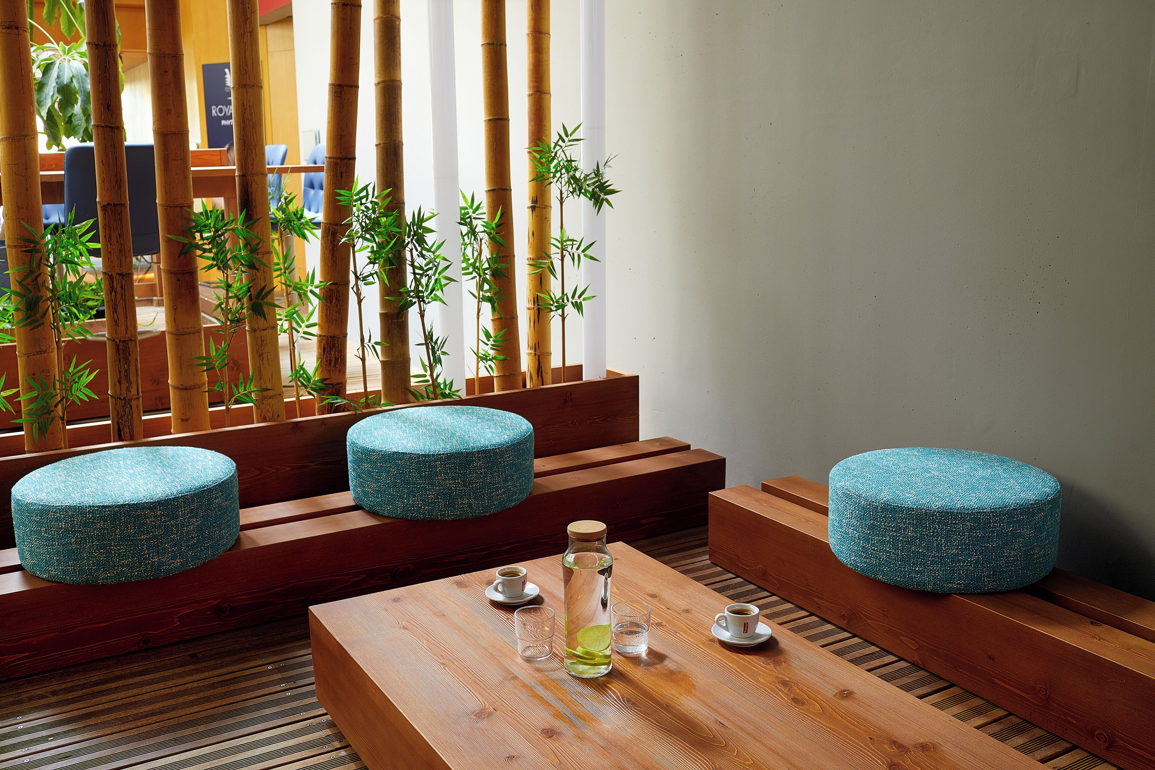 A modern seating area with wooden benches and round blue ottomans, a wooden table with a glass bottle and two glasses, and a bamboo screen in the background.