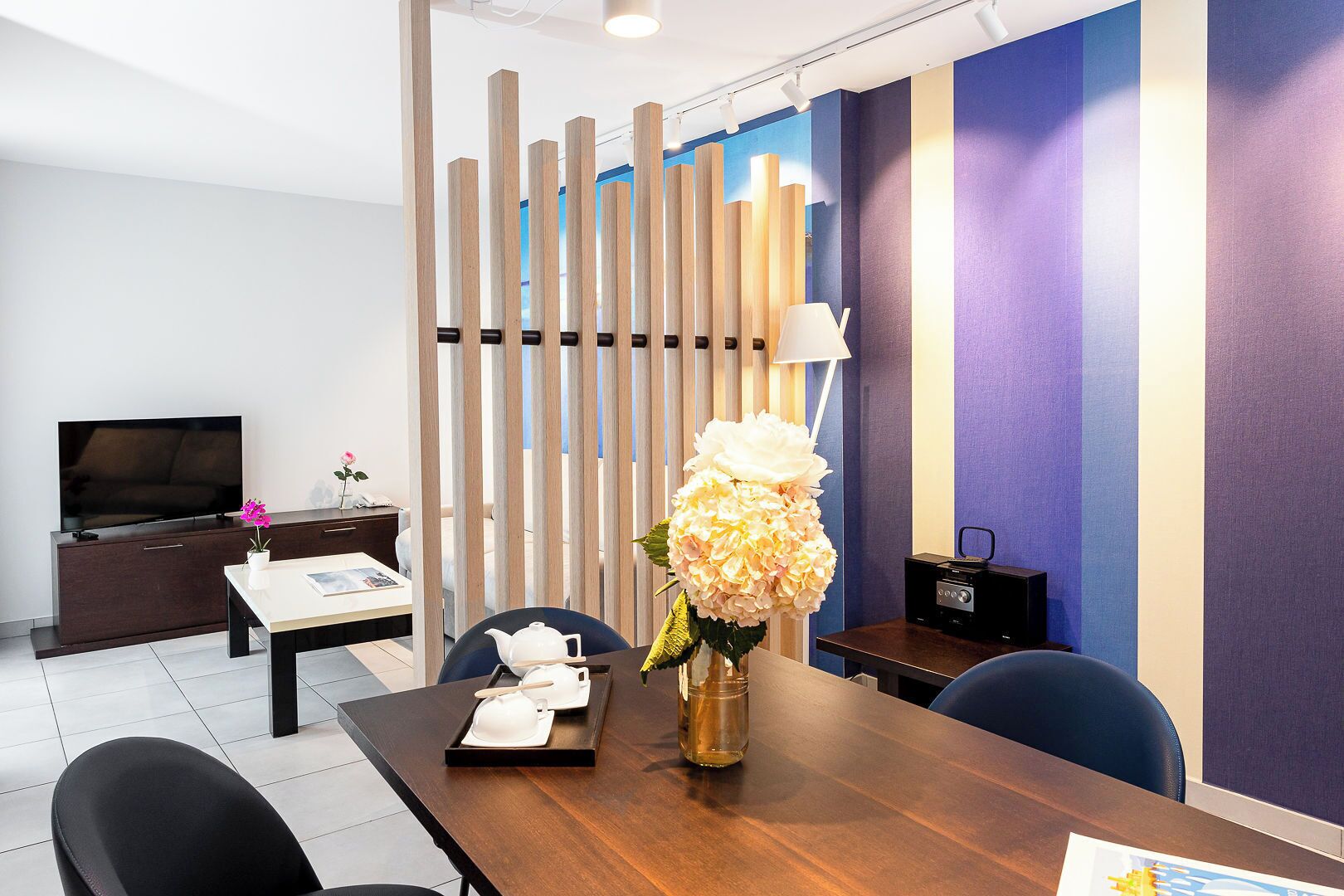 A modern hotel room with a wooden partition, a television, a desk with a lamp, and a dining area with a vase of flowers.