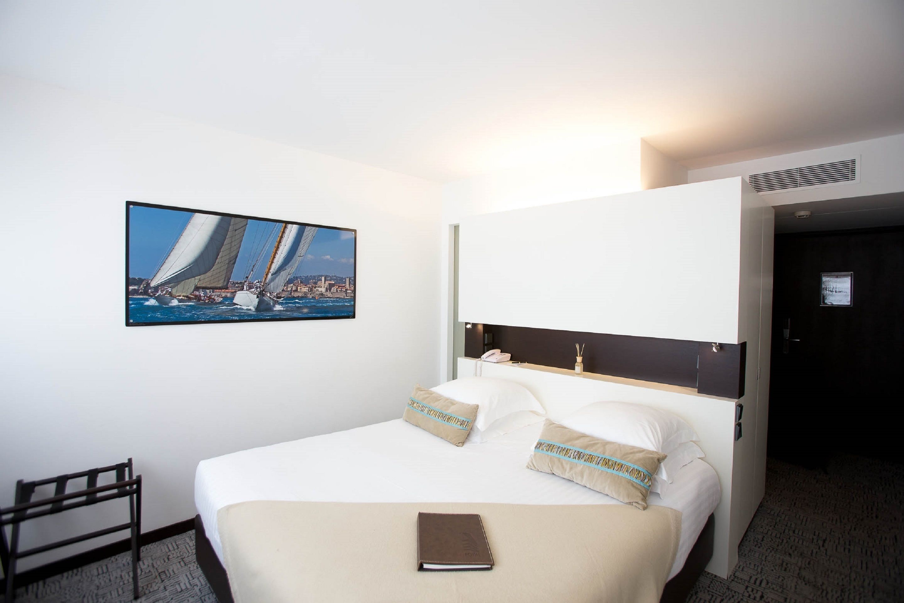 A hotel room with a bed, a bedside table, a chair, a painting of a sailboat, and a door.