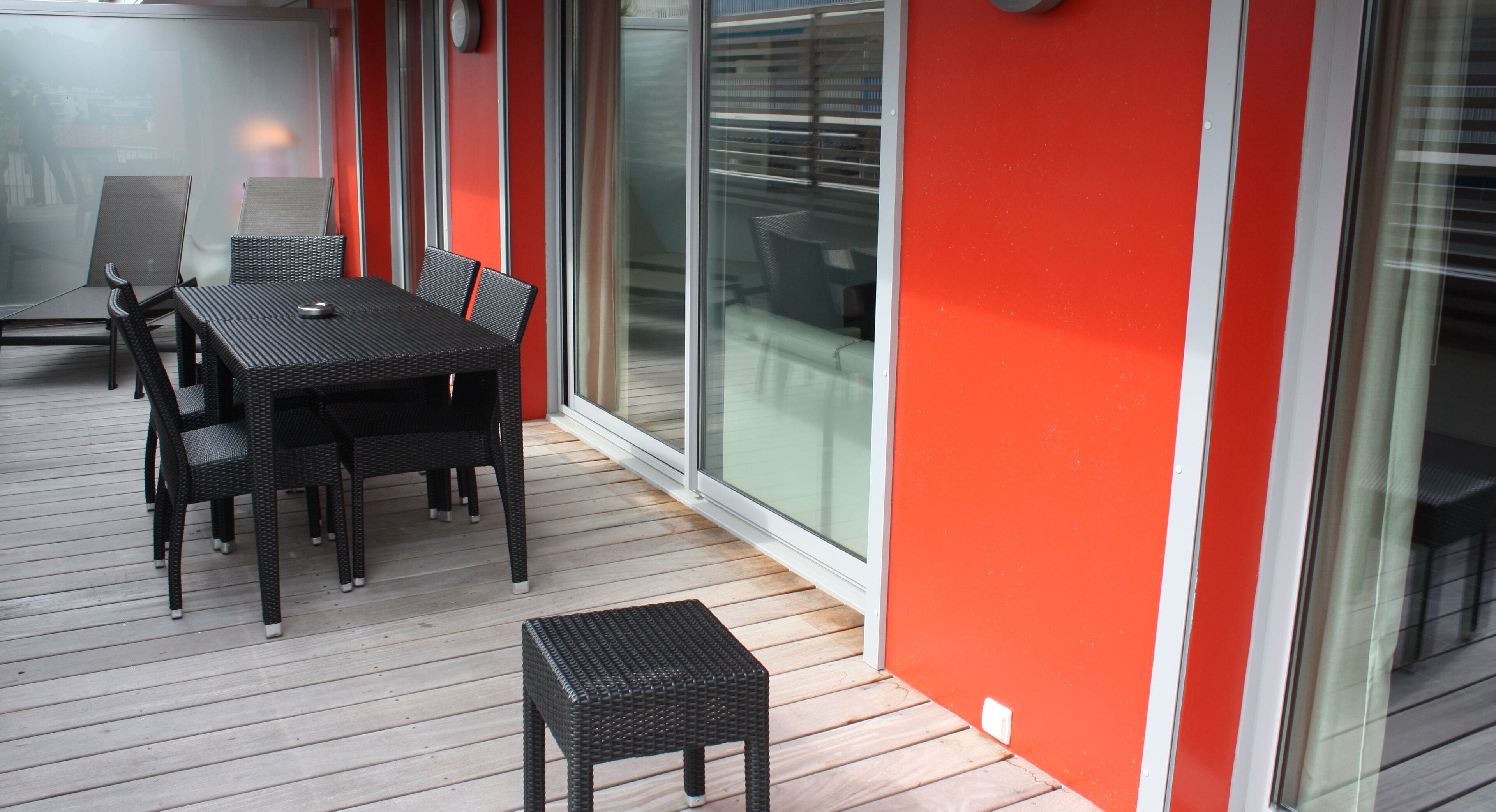Ein Essbereich mit Tisch und Stühlen auf einer Holzterrasse, neben einer roten Wand und großen Glas-Schiebetüren.