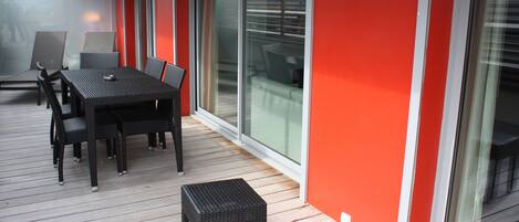 Un comedor con mesa y sillas sobre una terraza de madera, junto a una pared roja y grandes puertas correderas de vidrio.
