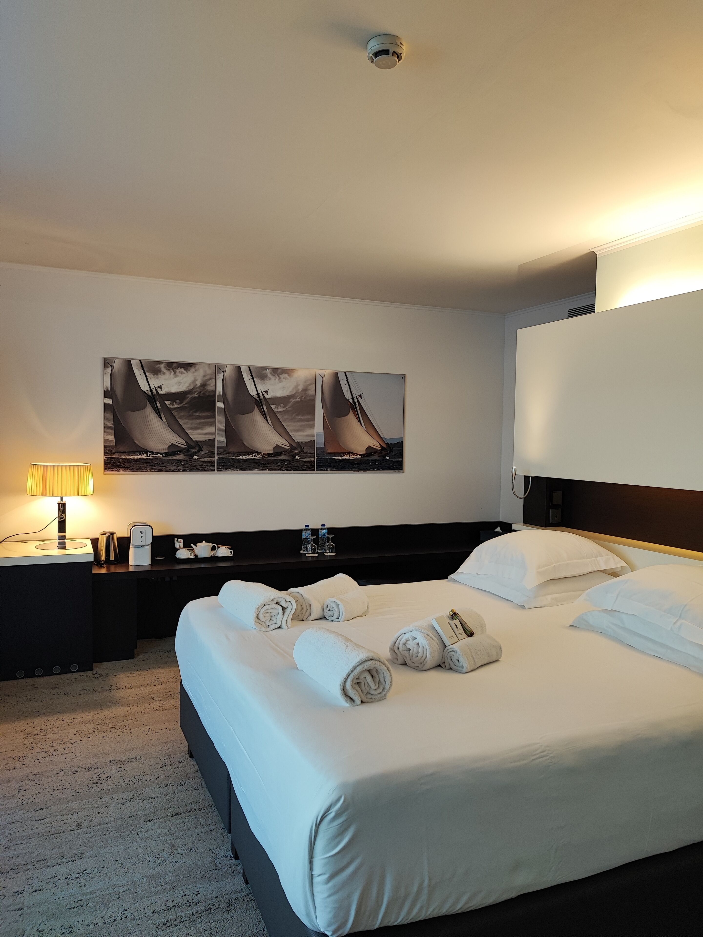 A hotel room with a large bed, a bedside table with a lamp, a coffee maker, and a painting of sailboats on the wall.