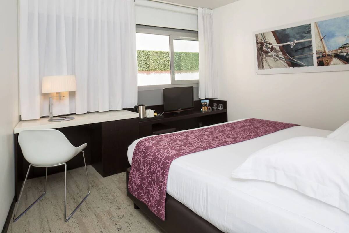 A hotel room with a bed, a desk with a chair, a television, and a window with curtains.