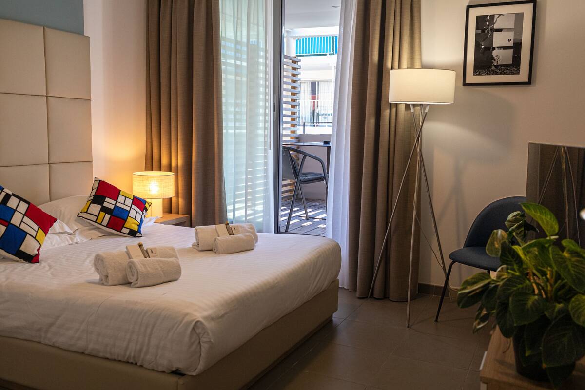 A hotel room with a bed, pillows, a bedside lamp, a chair, a potted plant, and a balcony with a view.