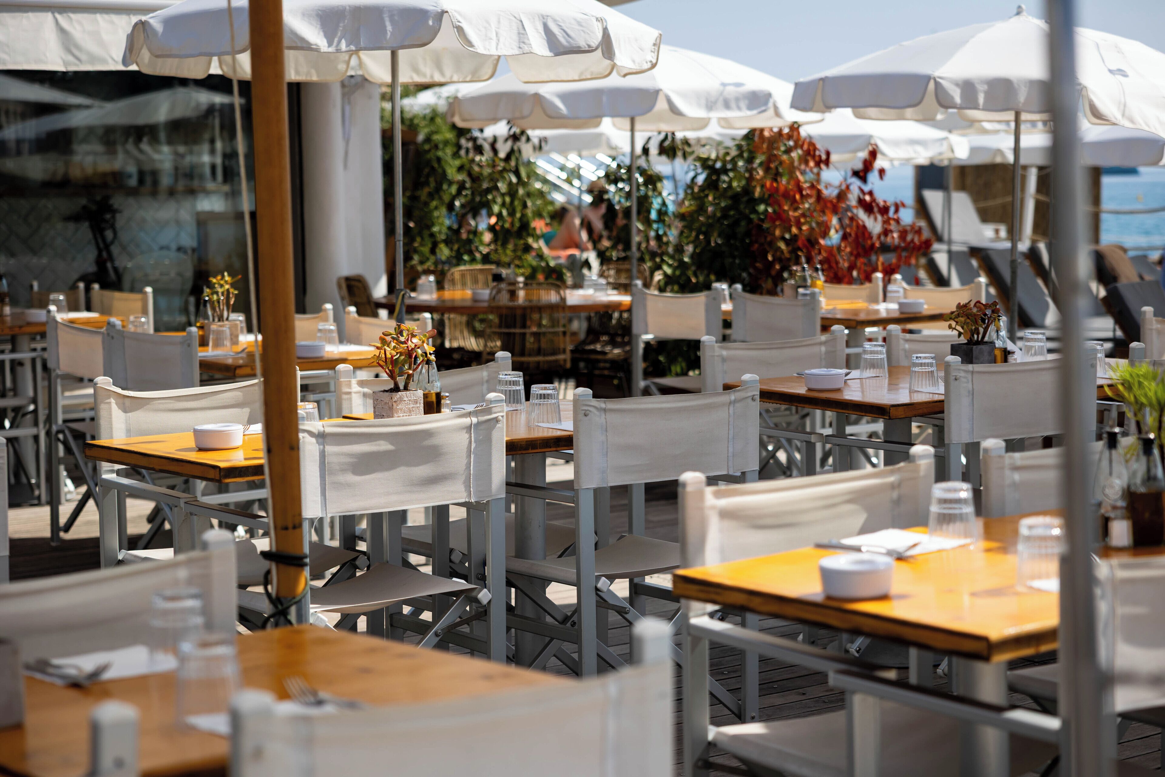 Outdoor tables and chairs arranged under white umbrellas, with a view of the sea and some greenery in the background.