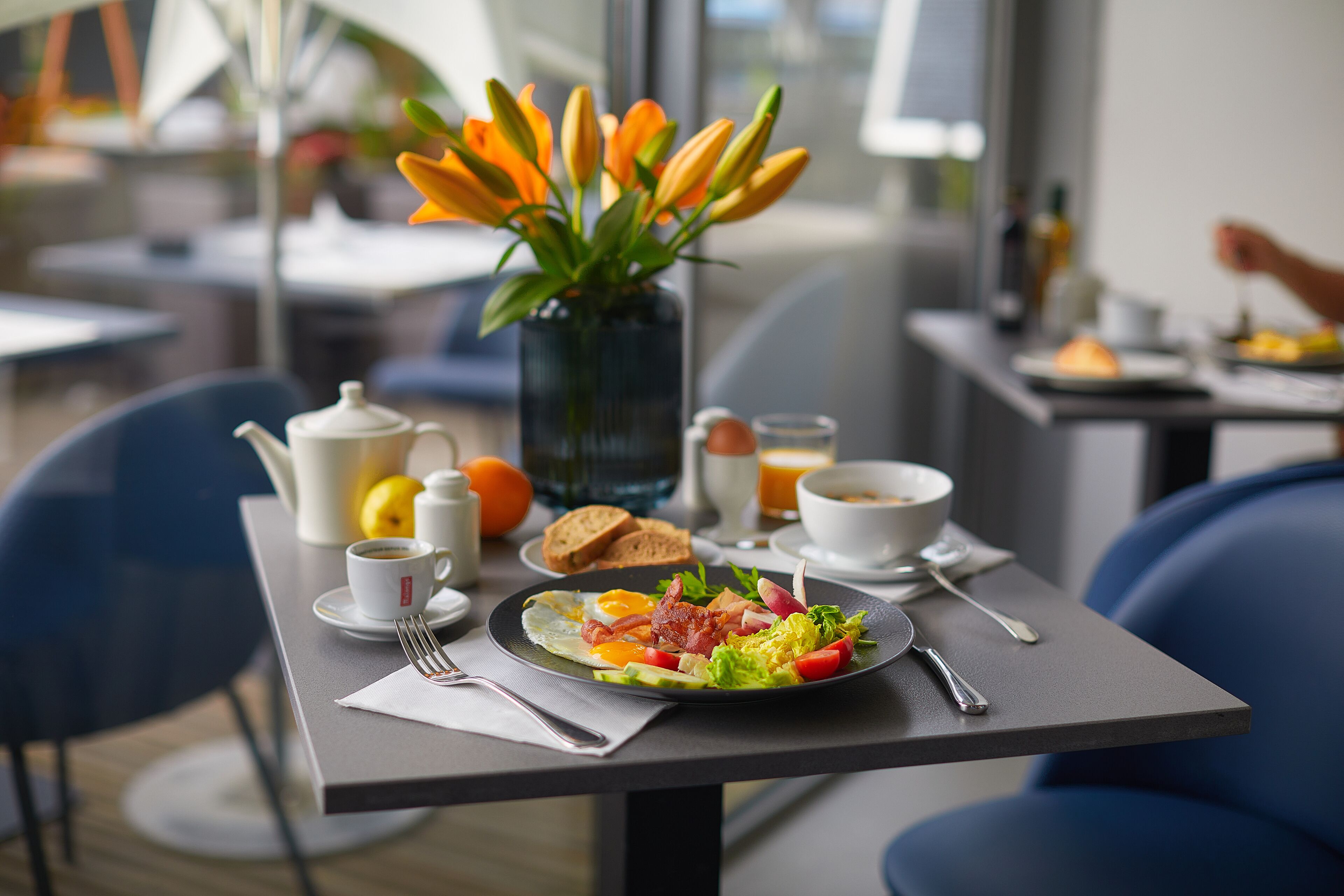 A breakfast table with a plate of eggs, bacon, and vegetables, a vase of flowers, and a coffee pot.