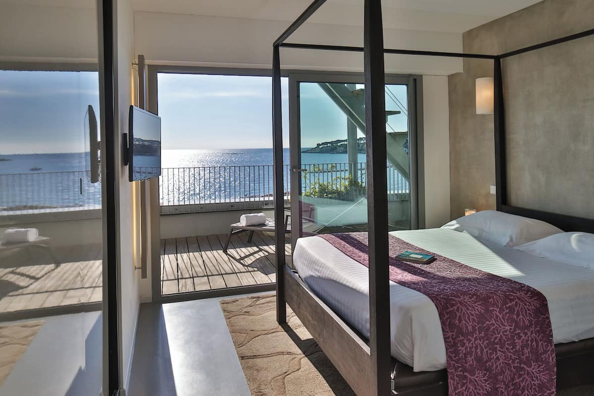 A modern hotel room with a balcony, a bed, a TV, and a view of the sea.