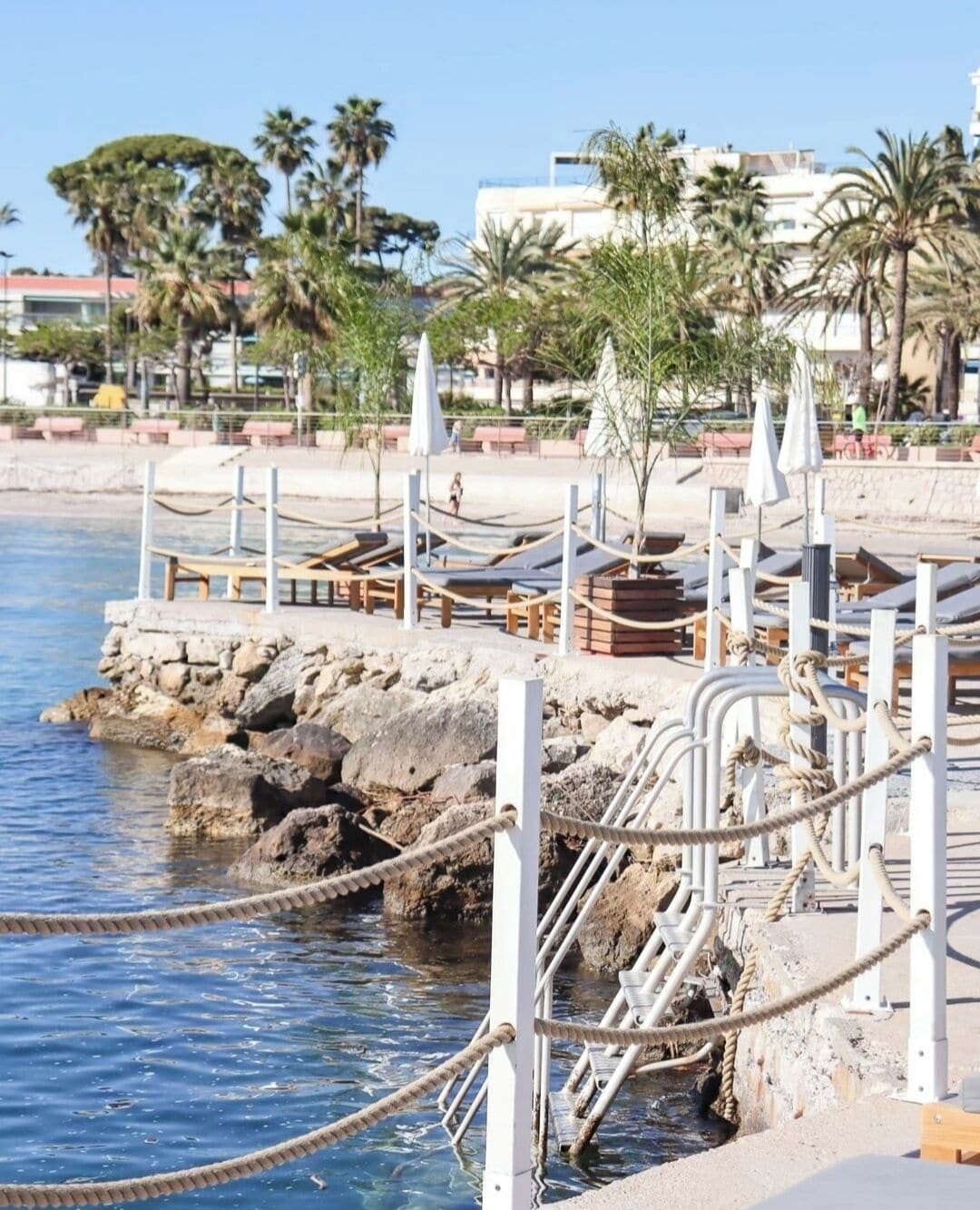 A coastal promenade with sun loungers, a rocky shoreline, and palm trees.