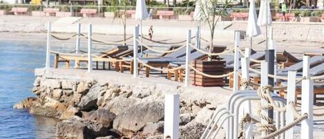 A coastal promenade with sun loungers, a rocky shoreline, and palm trees.