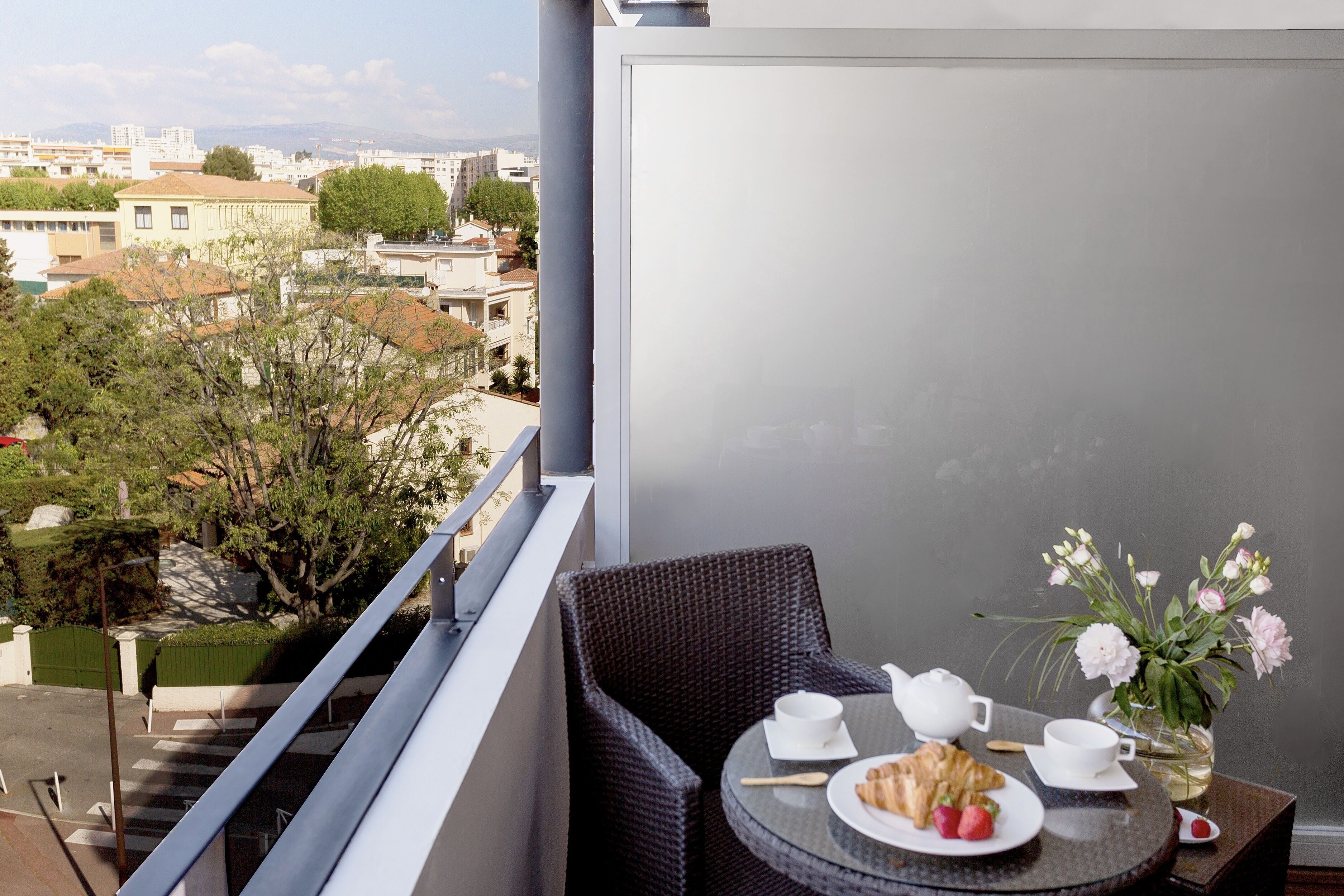 A balcony with a round table, a wicker chair, a vase with flowers, and a plate with food and a teapot.