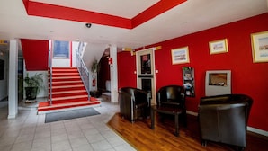 Un vestíbulo de hotel con paredes rojas, una escalera y suelo de madera. Se observan dos sillones de cuero y un mostrador de recepción.