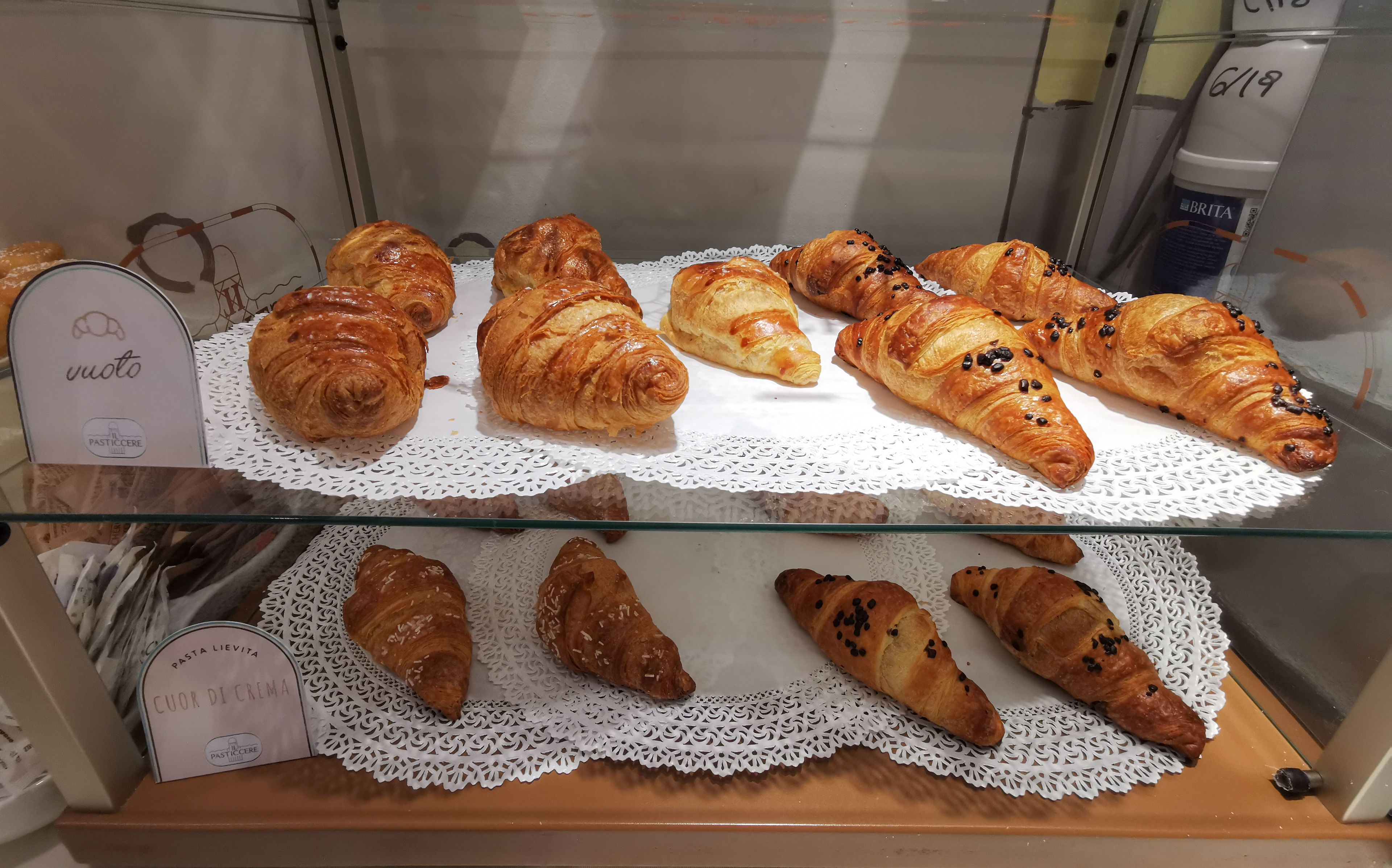 A display case with various types of pastries, including croissants and other baked goods, arranged on white doilies.