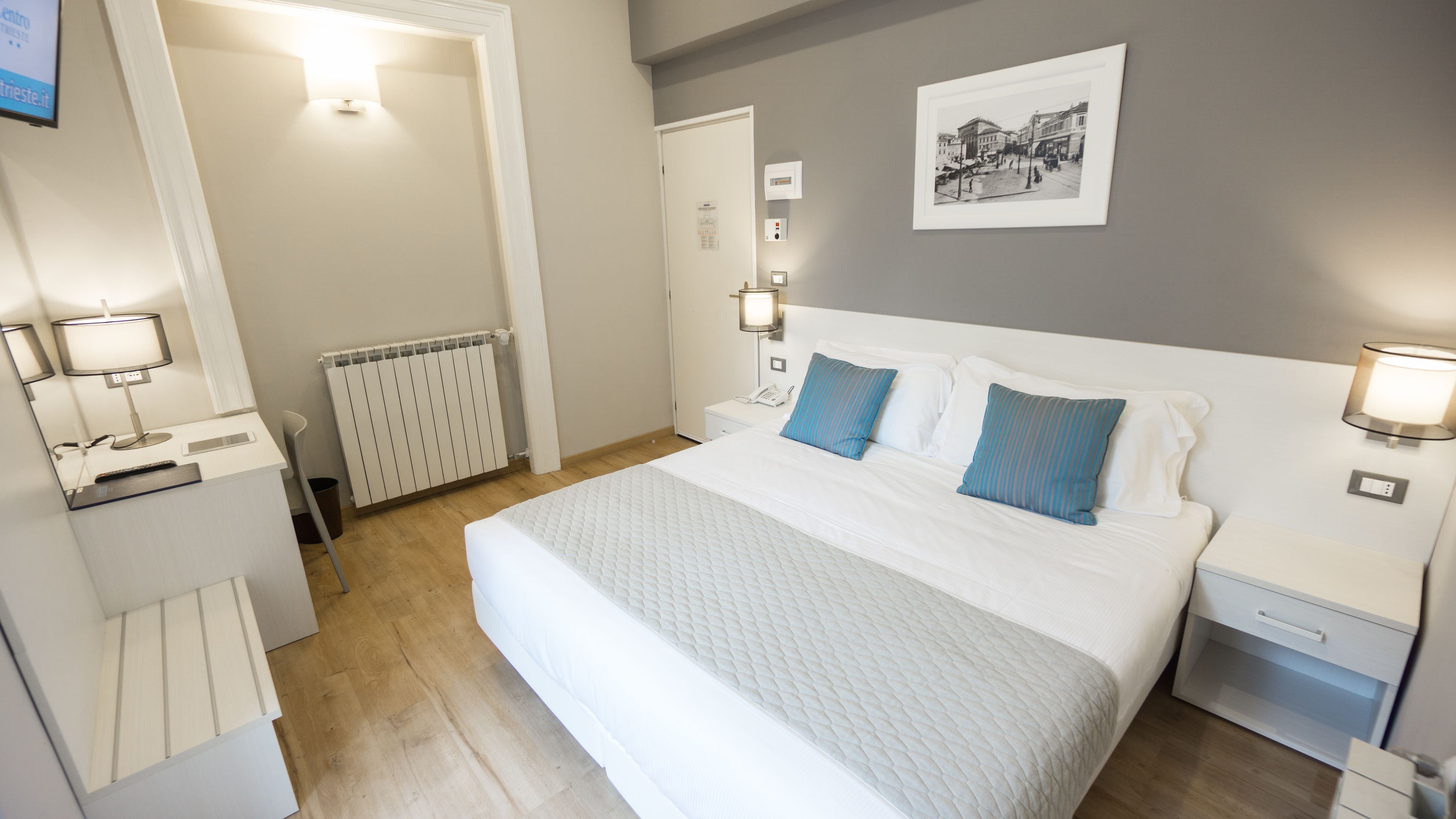 Habitación de hotel con una cama grande, lámparas de noche, un radiador, un escritorio con lámpara y un cuadro en la pared.