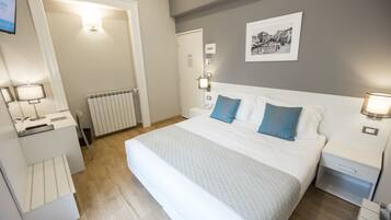 Habitación de hotel con una cama grande, lámparas de noche, un radiador, un escritorio con lámpara y un cuadro en la pared.