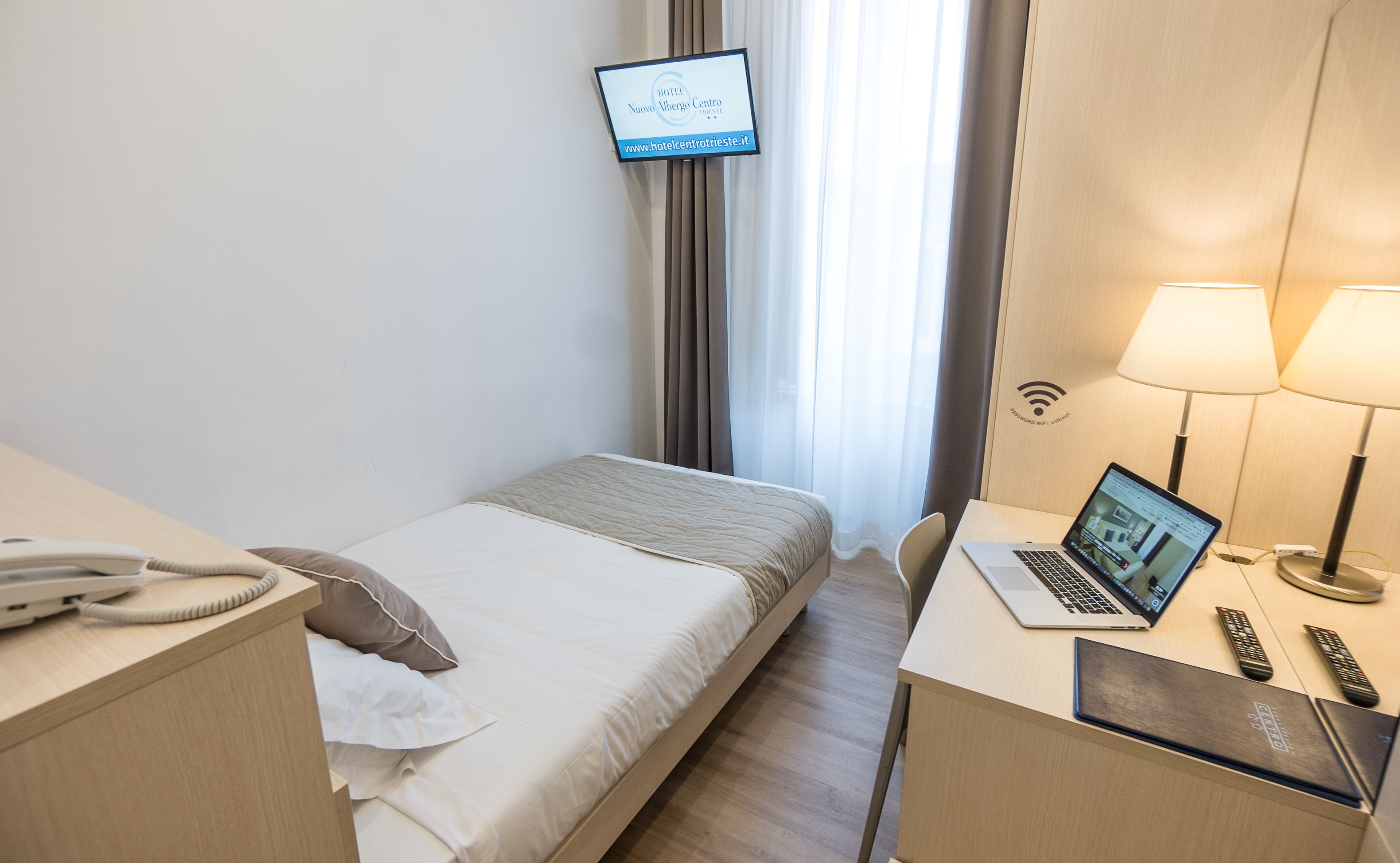 A hotel room with a bed, a desk with a laptop, a phone, and a TV displaying the hotel name.