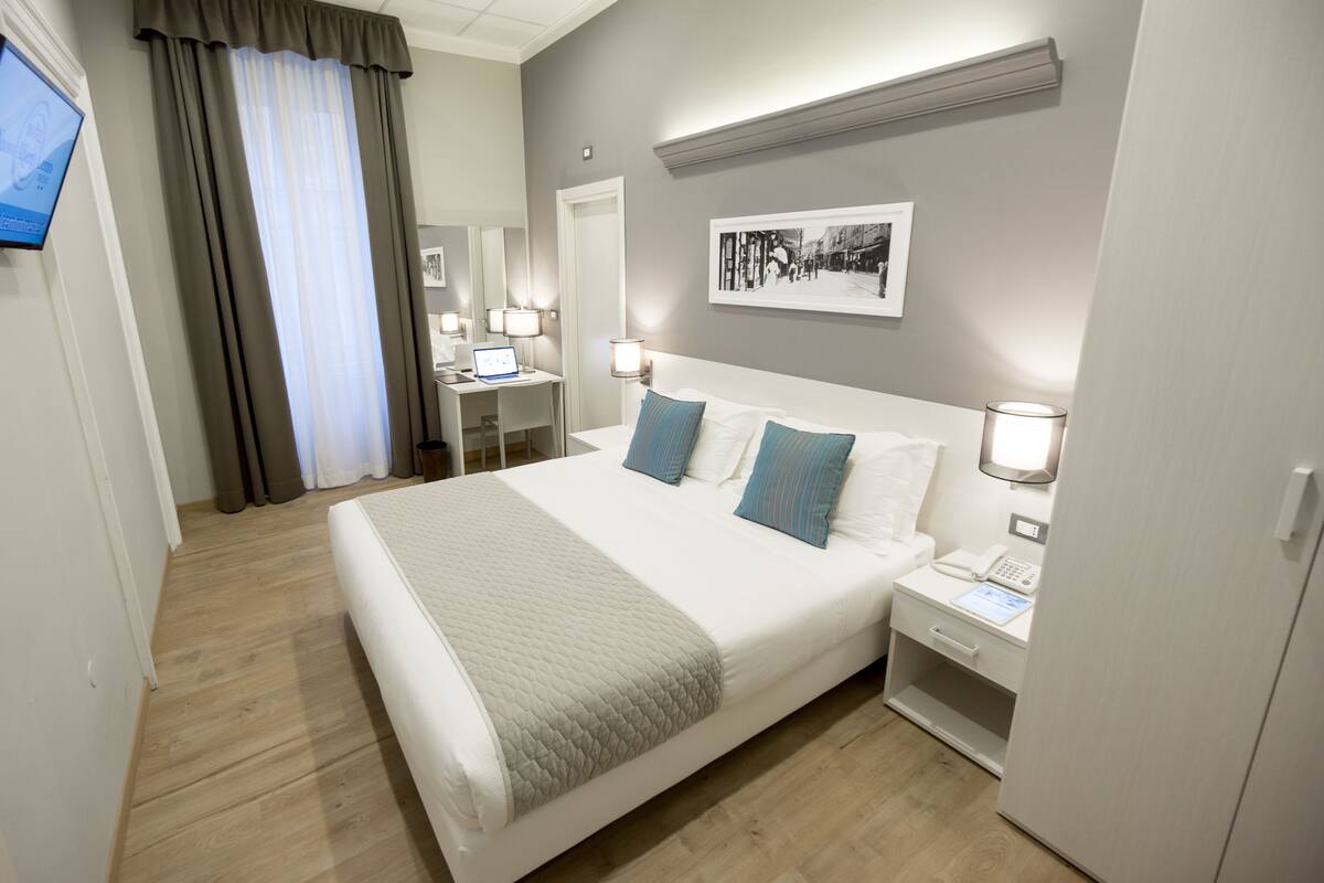 A hotel room with a large bed, bedside tables, a desk with a computer, and a flat-screen TV.