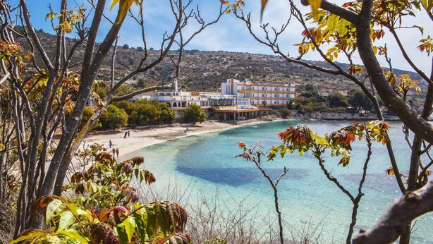 Un hotel frente al mar con vista a la playa, un edificio grande de varios pisos y un mar azul cristalino.