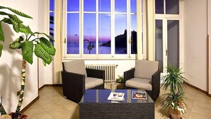 Salón con una mesa de centro de vidrio, dos sillones, un radiador y vistas al atardecer sobre el agua.